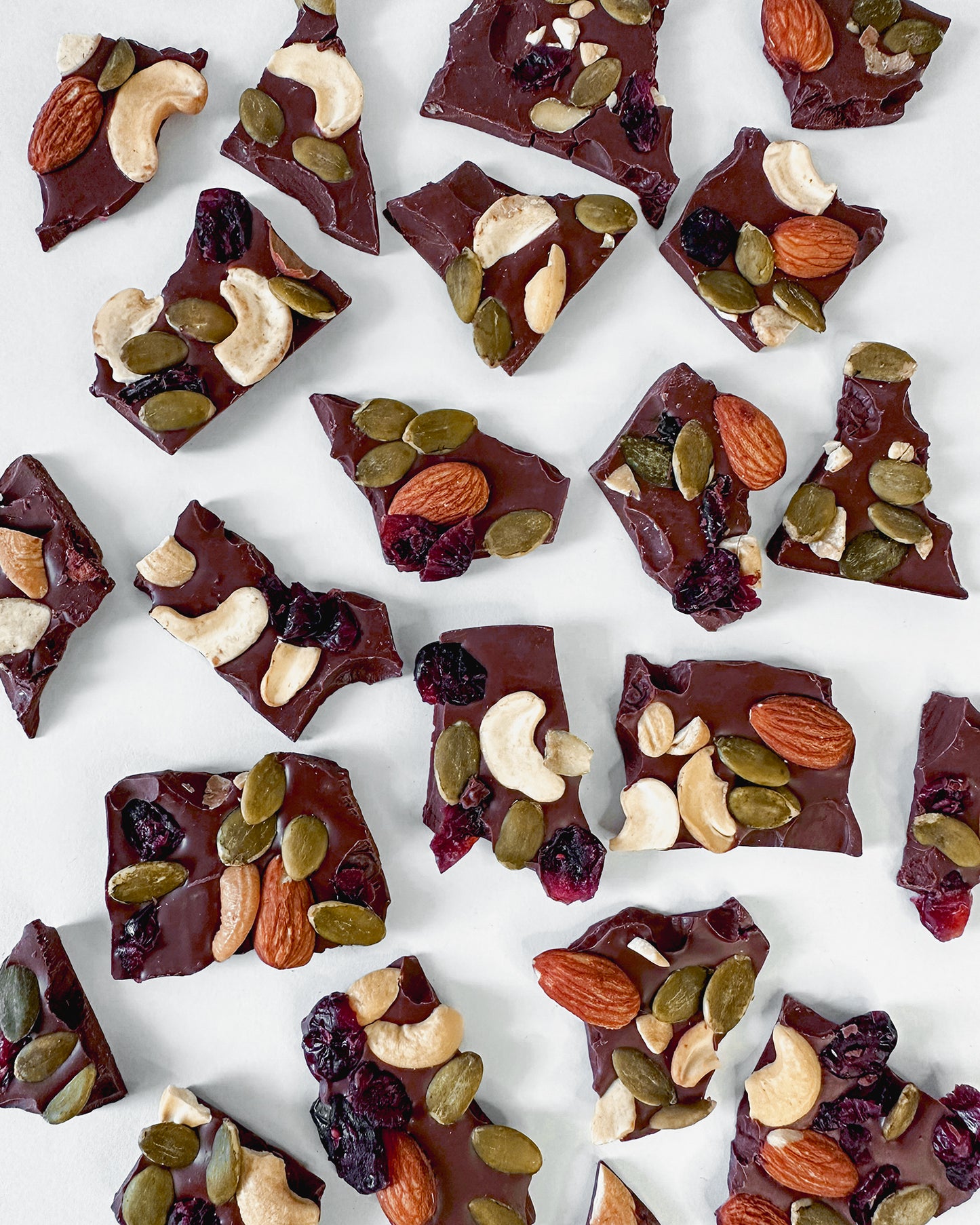 dark-milk-chocolate-barks-topped-with-trail-mix-made-of-almonds-cashews-pumpkin-seeds-dried-cranberries-scattered-on-white-surface